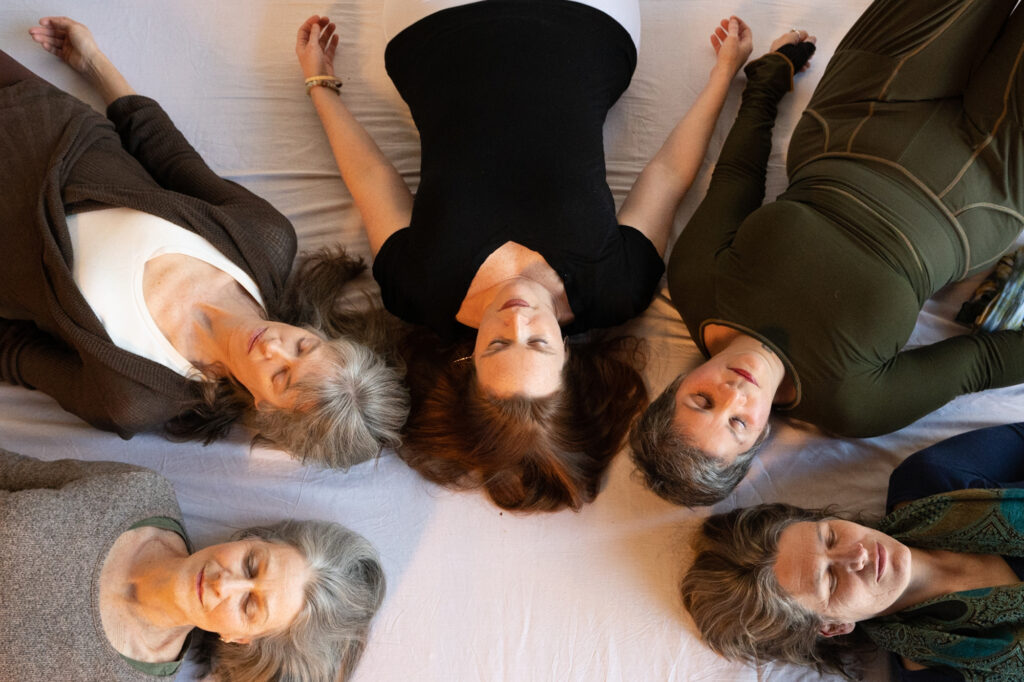 Five women lie on the floor in a circle with heads together in meditation